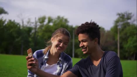 Cute-friends-resting-in-a-park,-sitting-on-the-grass,-looking-at-the-smartphone-and-laughing.-Mixed-race-couple-sitting-on-the