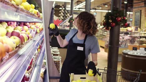 Side-view-of-pretty-caucasian-worker-in-black-apron-and-gloves-stocking-the-fruits-in-supermarket.-Young-employee-at-work.-Curly