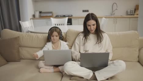 Mother-and-her-cute-little-girl-using-laptops-sitting-next-to-each-other-on-couch