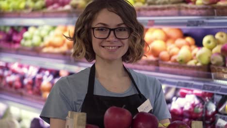 Schöne-Lächelnde-Junge-Supermarktangestellte-In-Schwarzer-Schürze-Hält-Eine-Kiste-Voller-Äpfel-Vor-Dem-Regal-Im-Supermarkt