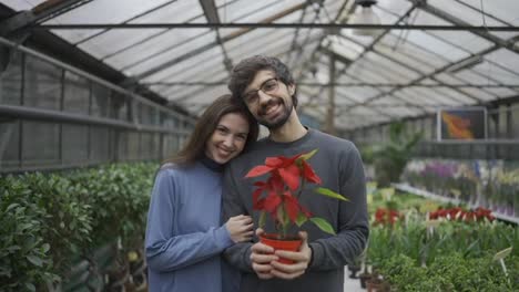 Beautiful-young-couple-in-casual-clothes-is-holding-a-plant,-looking-at-camera-and-smiling