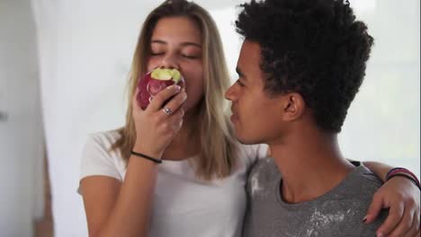 Young-multi-ethnic-couple-embracing-each-other-in-the-kitchen-while-cooking.-Beautiful-caucasian-girl-taking-the-apple-while-her