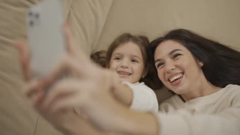 Mother-and-daughter-taking-selfie-at-home-while-lying-on-bed