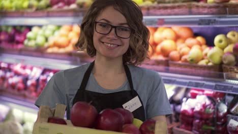Beautiful-smiling-young-female-supermarket-employee-in-black-apron-holding-a-box-full-of-apples-in-front-of-shelf-in-supermarket