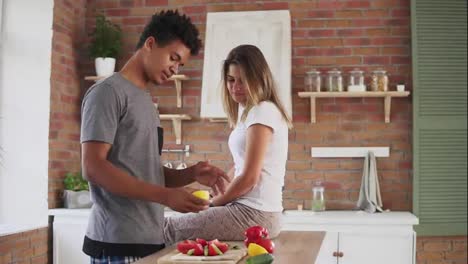Handsome-african-guy-preparing-salad,-cutting-lemon-and-squeezing-juice-from-it-in-a-funny-way-while-his-caucasian-girlfriend-is