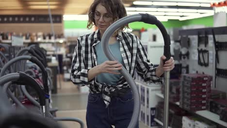 Pretty-curly-haired-woman-standing-near-new-vacuum-cleaners-in-home-appliance-store,-take-one-to-compare.-Various-vacuum