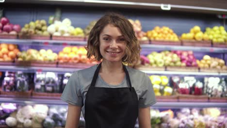 Portrait-of-attractive-young-saleswoman-in-black-apron-standing-in-supermarket-with-shelves-of-fruits-on-background,-looking-at
