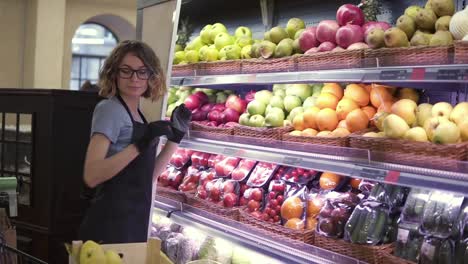 Close-up-of-pretty-caucasian-worker-in-black-apron-and-gloves-stocking-the-fruits-in-supermarket.-Young-employee-in-glasses-at