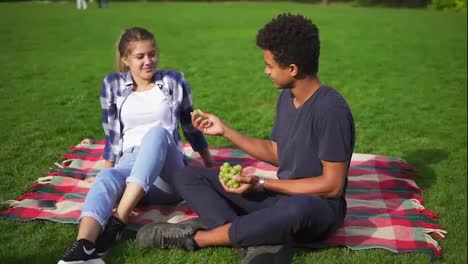 Young-lovely-couple-taking-fruit-sitting-on-the-grass-in-park-on-summer-day.-Man-carefully-feeds-woman-with-juicy-grapes
