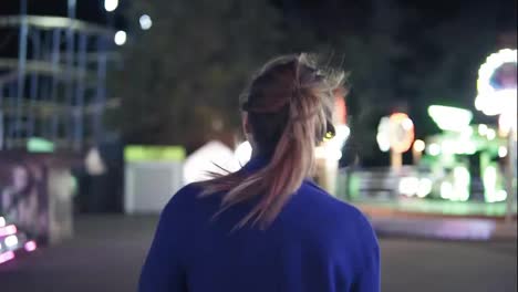 Young-smiling-girl-with-ponytail-walking-forward-then-turning-around-smiling-and-looking-at-camera-hanging-out-in-amusement-park