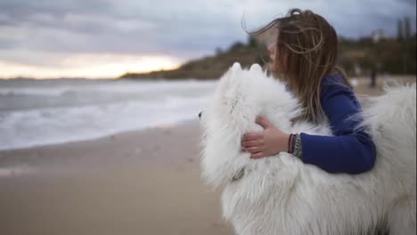 Side-view-of-a-young-woman-sitting-on-the-sand-and-embracing-her-dog-of-the-Samoyed-breed-by-the-sea.-White-fluffy-pet-on-the