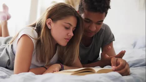 Attractive-african-guy-is-reading-a-book-out-loud-while-lying-in-bed-together-with-his-girlfriend-at-home-in-bedroom.-They-are