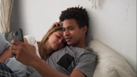 Young-beautiful-and-loving-multi-ethnic-couple-taking-selfie-picture-on-smartphone-camera-while-lying-in-bed-in-the-morning