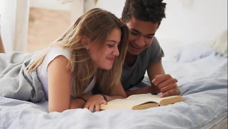 Happy-smiling-multi-ethnic-couple-lying-in-bed-reading-a-book-together-at-home-in-bedroom.-Slow-Motion-shot