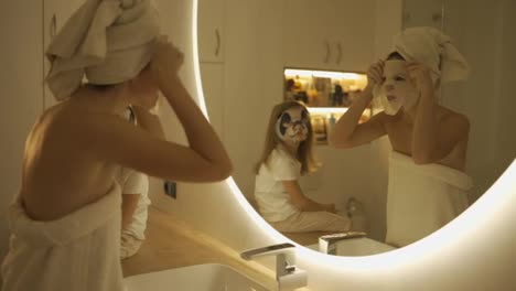 Mother-applying-tissue-panda-mask-on-her-face-relaxing-in-the-bedroom-with-her-daughter-next-to-her