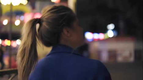 Back-view-of-young-attractive-woman-with-ponytail-walking-in-amusement-park-at-night,-turning-around-and-smiling-to-the-camera