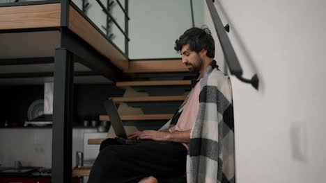 Young-bearded-caucasian-freelance-worker-use-laptop,-sitting-on-wooden-stairs-at-home-in-plaid