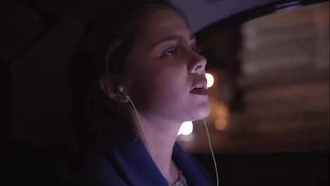 Beautiful-young-woman-is-riding-in-taxi-sitting-on-the-backseat-and-listening-to-the-music-in-earphones,-moving-her-hands