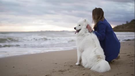 Back-view-of-a-young-woman-sitting-on-the-sand-and-embracing-her-dog-of-the-Samoyed-breed-by-the-sea.-White-fluffy-pet-on-the