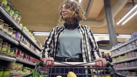 Cheerful-woman-in-plaid-shirt-and-headphones-on-neck-is-walking-in-grocery-store-steering-shopping-trolley-with-food-inside-it
