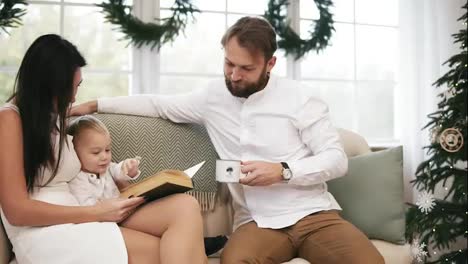 Young-mother-and-her-son-reading-book,-while-father-is-drinking-tea-from-his-cup,-listening-to-them.-Family-reading-Christmas