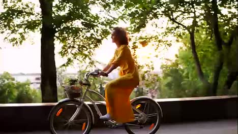 Brunette-smiling-woman-in-long-yellow-dress-is-enjoying-her-time-riding-a-city-bicycle-with-a-basket-and-flowers-inside-during