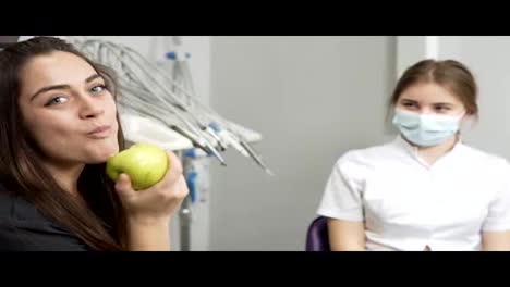 Young-woman-biting-a-green-apple-in-the-dental-office-while-sitting-near-the-dentist-after-the-procedures.-Showing-her-thumb-up