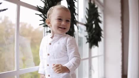 Cute-baby-boy-in-white-shirt-standing-on-the-window-sill-and-smiling.-The-window-is-decorated-with-Christmas-wreath