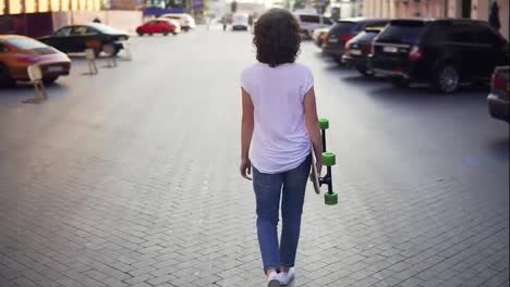 Back-view-of-a-woman-walking-in-the-old-city-street-holding-her-longboard-in-the-morning,-parking-in-the-city.-Beautiful-old