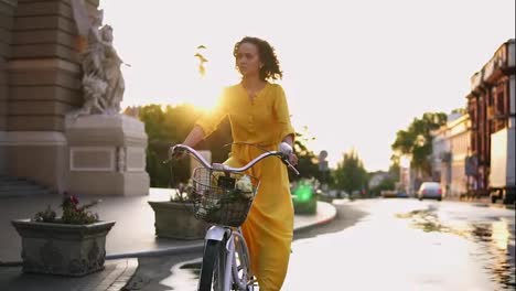 Beautiful-woman-riding-a-city-bicycle-with-a-basket-and-flowers-in-the-city-center-during-the-dawn-enjoying-her-time-early-in