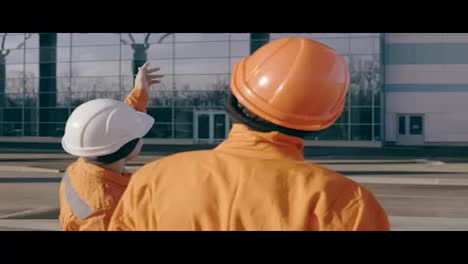 Zwei-Arbeiter-In-Orangefarbener-Uniform-Und-Helm-Zeigen-Auf-Das-Gebäude-Und-Untersuchen-Das-Objekt.-Zeitlupe