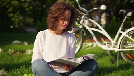 Young-woman-in-white-shirt-and-blue-jeans-is-reading-a-book-sitting-on-the-grass-in-park-and-drinking-her-green-detox-smoothie