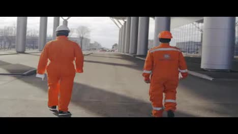 Two-happy-construction-workers-in-orange-uniform-and-helmets-walking-and-jumping-together
