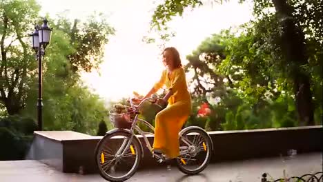 Beautiful-woman-in-long-yellow-dress-riding-a-city-bicycle-with-a-basket-and-flowers-inside-during-the-dawn.-Lens-flare