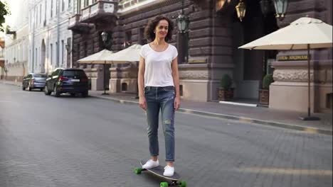 Attractive-brunette-woman-in-white-t-shirt,-blue-jeans-and-white-sneakers-skateboarding-at-sunrise-in-the-city-street-early-in