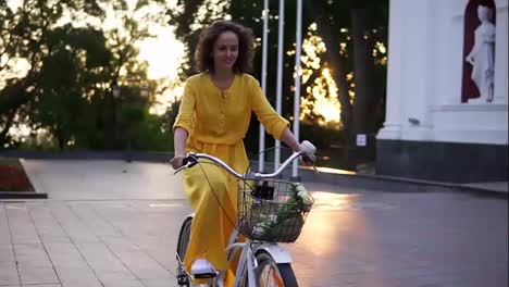 Young-attractive-woman-is-riding-a-city-bicycle-with-a-basket-and-flowers-in-the-city-park-while-the-son-is-raising.-Lens-flare