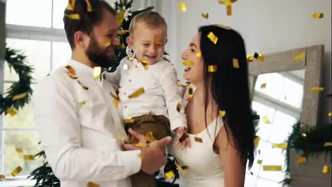Young-mother,-father-and-their-little-son-celebrating-with-shooting-confetti-on-Christmas-Eve-at-home,-kissing-their-cute-child