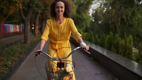 Beautiful-woman-in-long-yellow-dress-riding-a-city-bicycle-with-a-basket-and-flowers-in-the-city-park-looking-in-the-camera-and