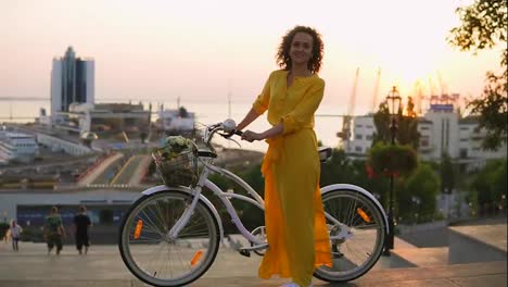 Young-brunette-woman-in-long-yellow-dress-standing-by-her-city-bike-holding-its-handlebar-with-flowers-in-its-basket-during-the