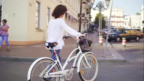 Back-view-of-a-brunette-woman-in-a-white-shirt-and-blue-jeans-walking-in-the-city-street-holding-her-bicycle-handlebar-with-a