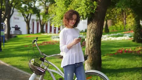 Portrait-of-a-young-woman-typing-a-message-using-her-smartphone-standing-in-the-city-park-near-her-city-bicycle-with-flowers-in