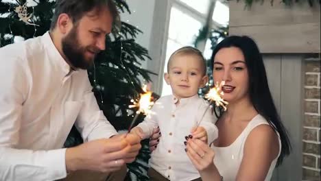 Happy-family-and-Bengal-fire-at-home-by-the-Christmas-tree.-Mother-and-father-holding-sparkler,-while-their-cute-child-is