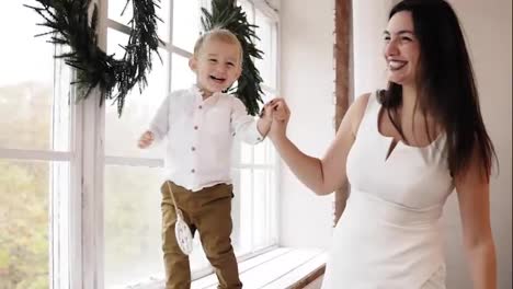 Young-smiling-mother-in-white-dress-is-holding-her-son's-hand-while-he-is-walking-on-the-window-sill-decorated-with-Christmas