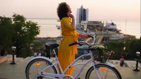 Attractive-brunette-woman-in-a-long-yellow-dress-during-the-dawn-standing-by-her-city-bicycle-holding-its-handlebar-with-flowers
