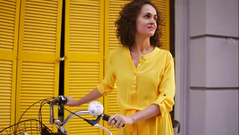 Happy-brunette-woman-in-a-long-yellow-dress-standing-by-the-yellow-wooden-fence-holding-her-city-bicycle-handlebar-with