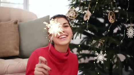 Smiling-friendly-attractive-young-woman-sitting-on-the-floor-by-the-Christmas-tree-celebrating-Xmas-with-sparklers-and-looking