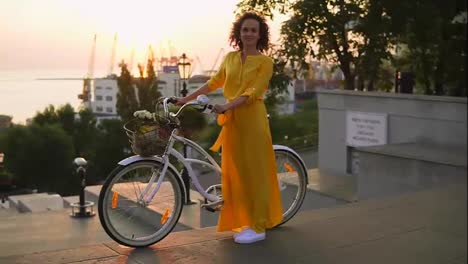 Smiling-brunette-woman-in-a-long-yellow-dress-standing-by-her-city-bicycle-holding-its-handlebar-with-flowers-in-its-basket