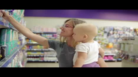 Young-attractive-mother-in-glasses-holding-her-child-in-her-arms-while-choosing-diapers-on-the-shelves-in-the-supermarket
