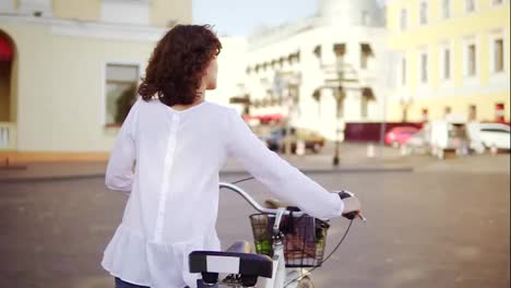 Back-view-of-a-woman-in-a-white-shirt-and-blue-jeans-walking-in-the-city-street-holding-her-bicycle-handlebar-with-a-basket