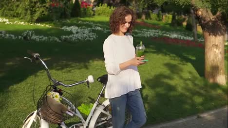 Portrait-of-a-young-woman-surfing-in-the-internet-using-her-smartphone-standing-in-the-city-park-near-her-city-bicycle-with
